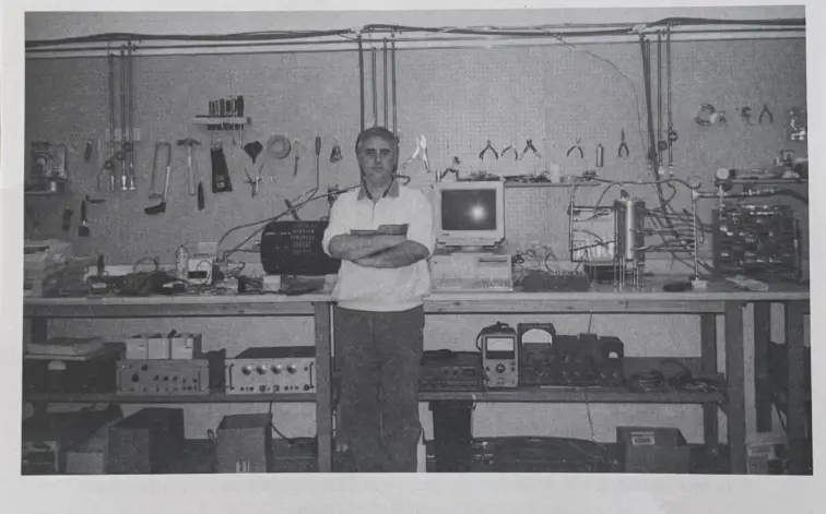 Dr. Kiril Chukanov in his Energy Research Laboratory Black and white photograph of dr. Kiril chukanov standing with his arms crossed in his laboratory, surrounded by workbenches equipped with testing electronics, hand tools, and a computer monitor.